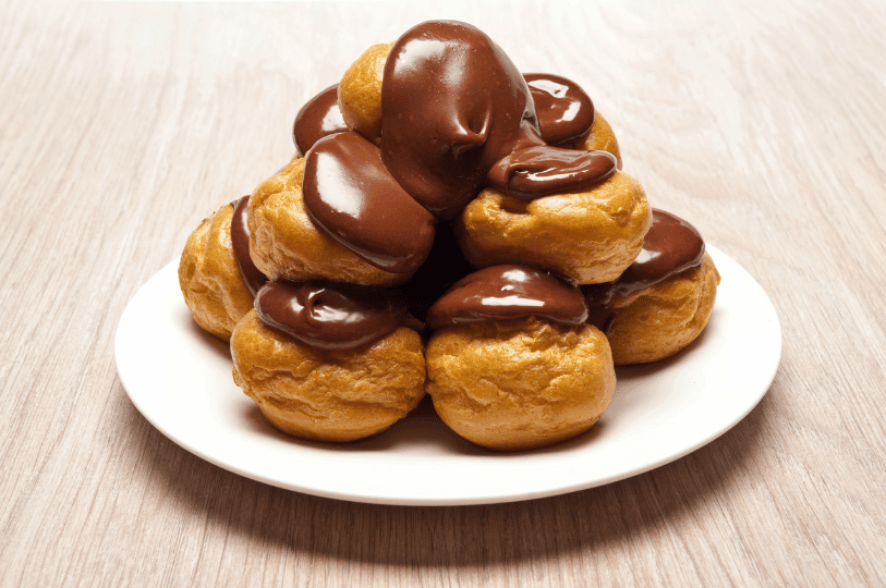 A neatly stacked pyramid of golden profiteroles served on a round white plate, cascading with a thick, glossy chocolate sauce. A neatly stacked pyramid of golden profiteroles served on a round white plate, cascading with a thick, glossy chocolate sauce.