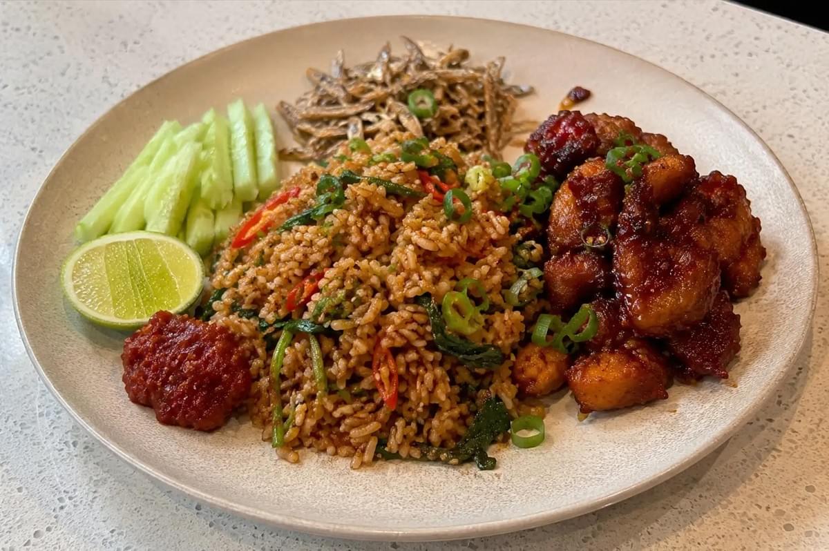 A large round ceramic plate features a mound of Malay-style kampung fried rice stir-fried with green leafy vegetables and sliced red chilies. Surrounding the rice are distinct side offerings: a pile of sticky, dark red sambal chicken, a heap of crispy fried anchovies (ikan bilis), fresh cucumber spears, a lime wedge, and a dollop of vibrant red sambal paste. A large round ceramic plate features a mound of Malay-style kampung fried rice stir-fried with green leafy vegetables and sliced red chilies. Surrounding the rice are distinct side offerings: a pile of sticky, dark red sambal chicken, a heap of crispy fried anchovies (ikan bilis), fresh cucumber spears, a lime wedge, and a dollop of vibrant red sambal paste.