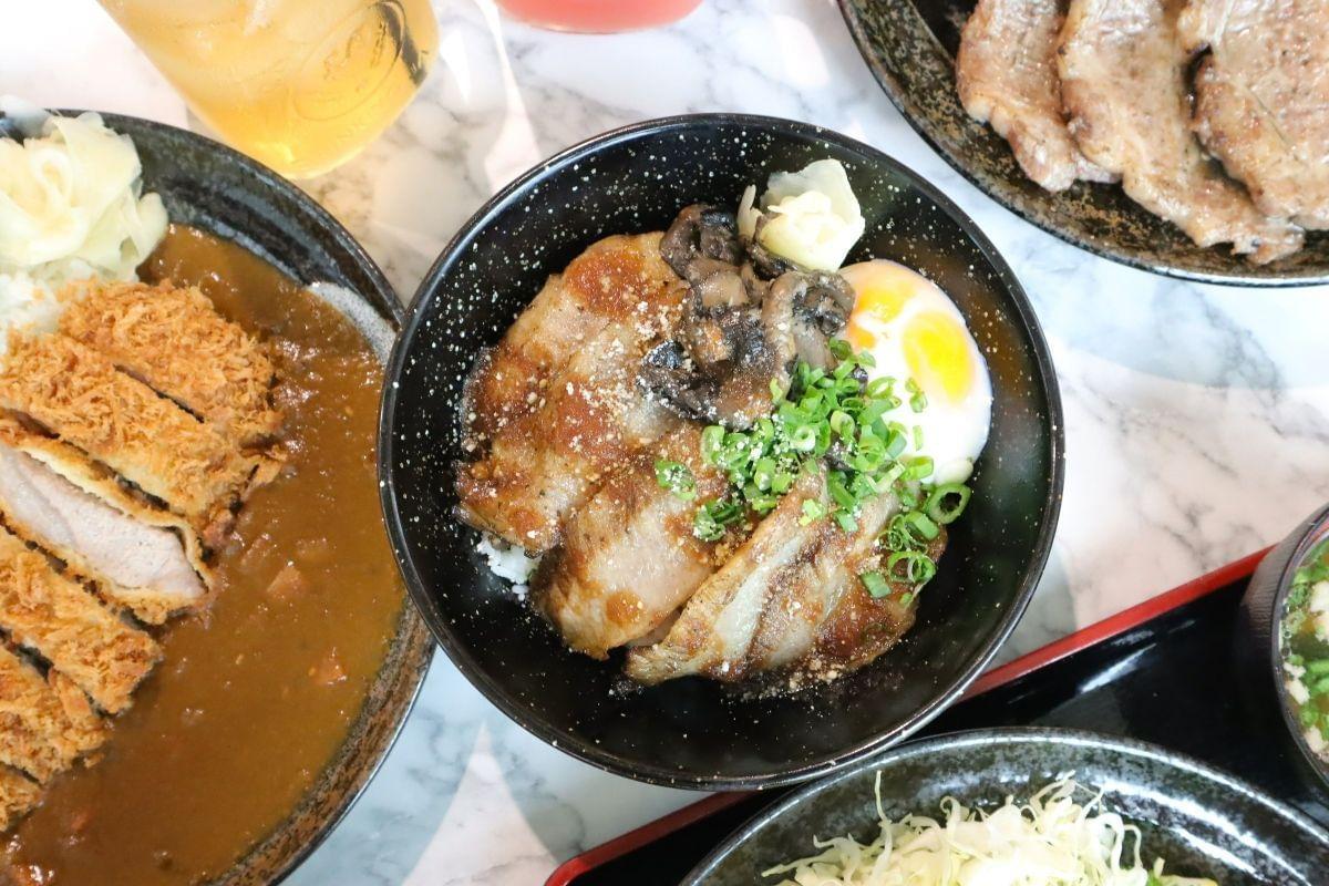 A top-down view features a Japanese rice bowl topped with seared pork belly, a soft-boiled egg, mushrooms, and chopped green onions. The dish is surrounded by other plates on a marble table, including pork katsu curry and grilled meat. A top-down view features a Japanese rice bowl topped with seared pork belly, a soft-boiled egg, mushrooms, and chopped green onions. The dish is surrounded by other plates on a marble table, including pork katsu curry and grilled meat.