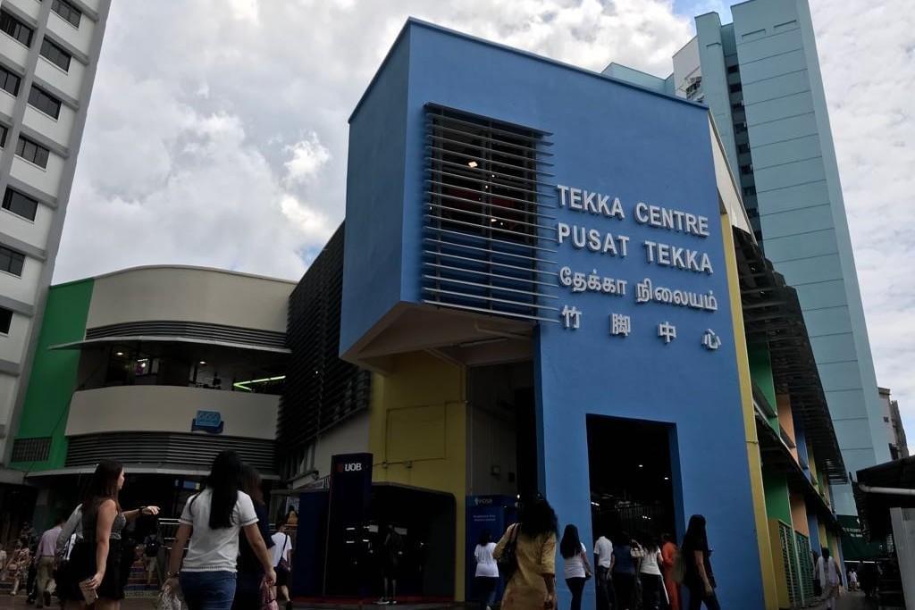 A bright blue architectural facade prominently displays the name "TEKKA CENTRE" in English, Malay, Tamil, and Chinese script. Pedestrians walk along the street level below the modern entrance structure on a partly cloudy day. A bright blue architectural facade prominently displays the name "TEKKA CENTRE" in English, Malay, Tamil, and Chinese script. Pedestrians walk along the street level below the modern entrance structure on a partly cloudy day.