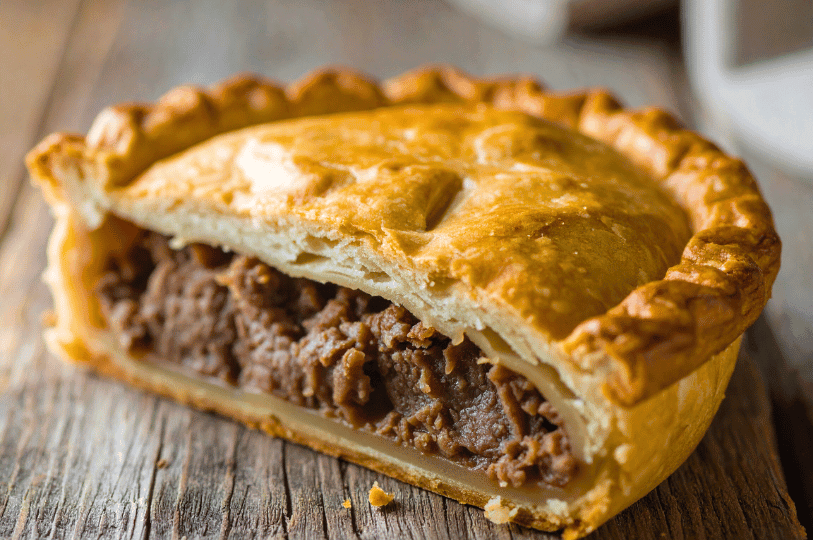 A cross-section of a golden-brown Australian meat pie showing a thick, savory minced beef filling inside a flaky shortcrust base and puff pastry lid. A cross-section of a golden-brown Australian meat pie showing a thick, savory minced beef filling inside a flaky shortcrust base and puff pastry lid.