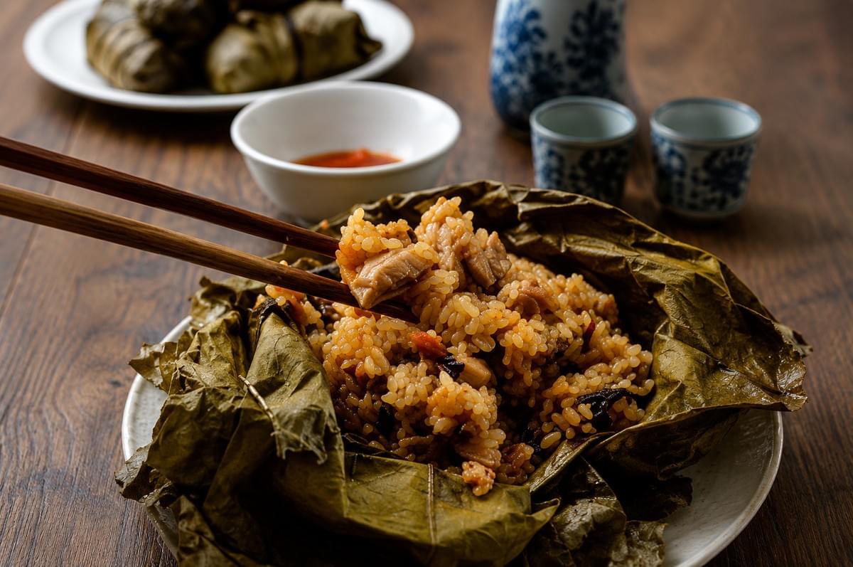 A close-up shot of seasoned sticky rice mixed with meat, served open on a large, dried lotus leaf, with wooden chopsticks lifting a bite. A close-up shot of seasoned sticky rice mixed with meat, served open on a large, dried lotus leaf, with wooden chopsticks lifting a bite.