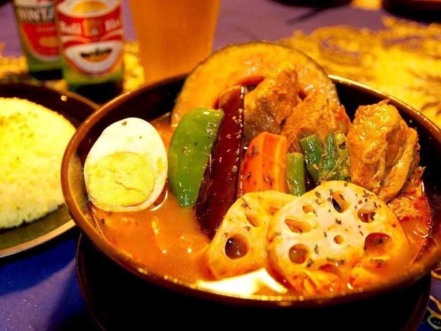 A large bowl of vibrant Japanese soup curry is filled with tender meat, a hard-boiled egg, and assorted vegetables like lotus root, green pepper, and eggplant. In the background, a plate of yellow rice and a glass of beer sit on a blue tablecloth. A large bowl of vibrant Japanese soup curry is filled with tender meat, a hard-boiled egg, and assorted vegetables like lotus root, green pepper, and eggplant. In the background, a plate of yellow rice and a glass of beer sit on a blue tablecloth.