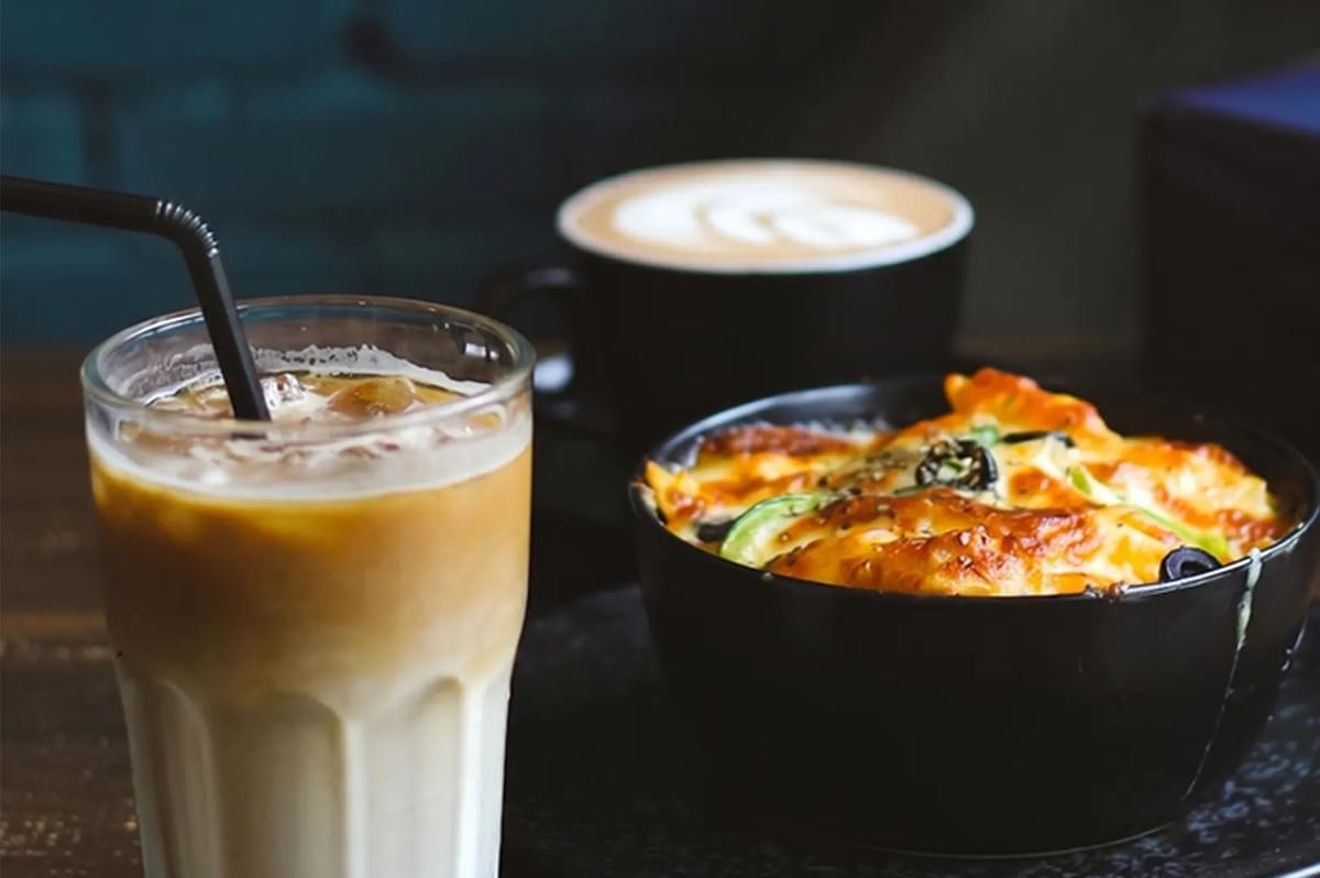 Iced coffee latte served with a baked vegetable pasta dish topped with melted cheese and olives, accompanied by a hot cappuccino in the background. Iced coffee latte served with a baked vegetable pasta dish topped with melted cheese and olives, accompanied by a hot cappuccino in the background.