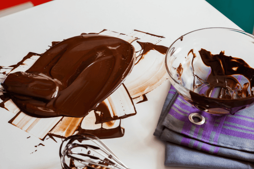 Melted chocolate spread out on a white stone work surface, sitting next to a partially scraped glass bowl and a wire whisk on a folded kitchen towel. Melted chocolate spread out on a white stone work surface, sitting next to a partially scraped glass bowl and a wire whisk on a folded kitchen towel.