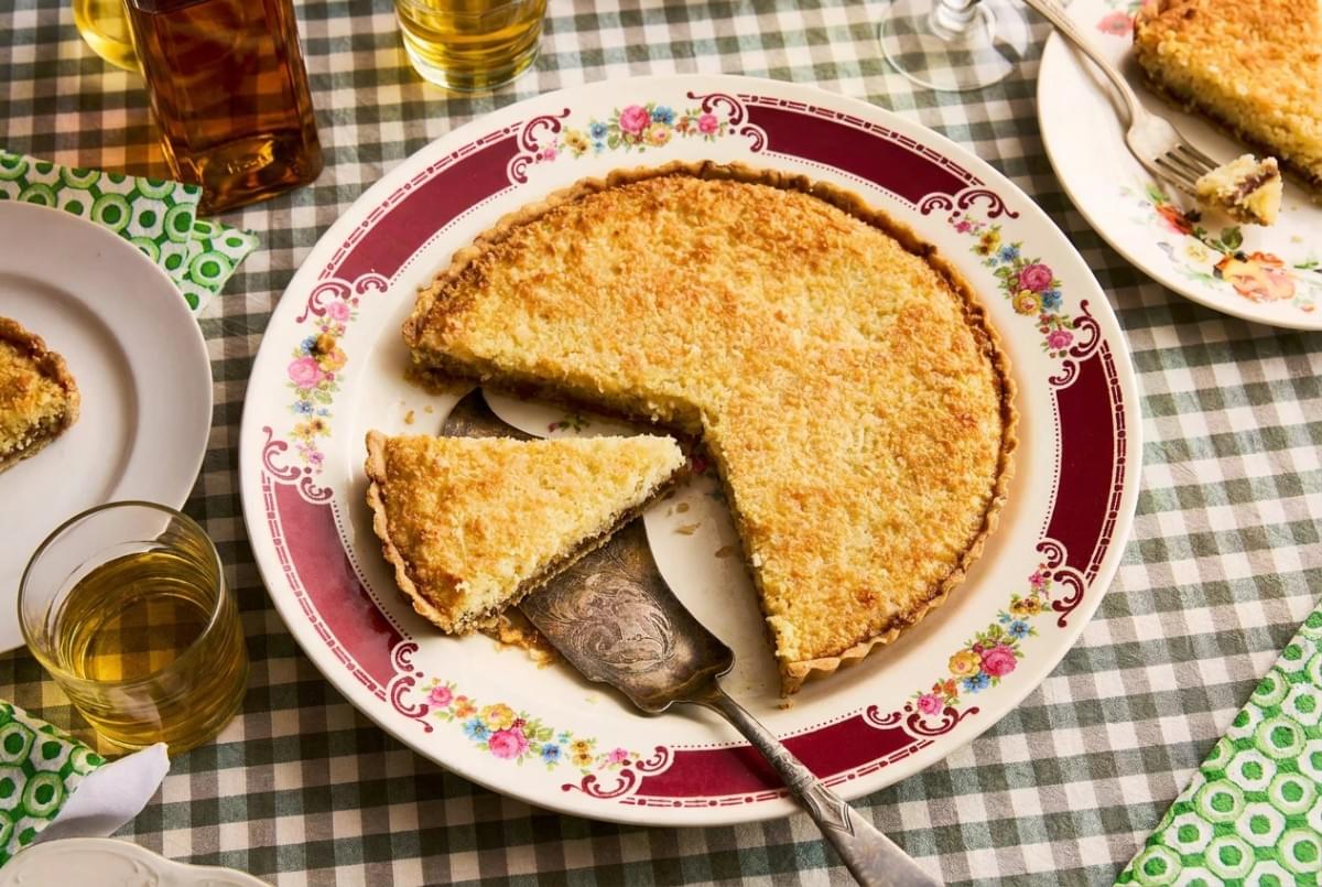 Golden coconut tart on a floral pattern plate with a pie server lifting one slice on a checkered tablecloth. Golden coconut tart on a floral pattern plate with a pie server lifting one slice on a checkered tablecloth.