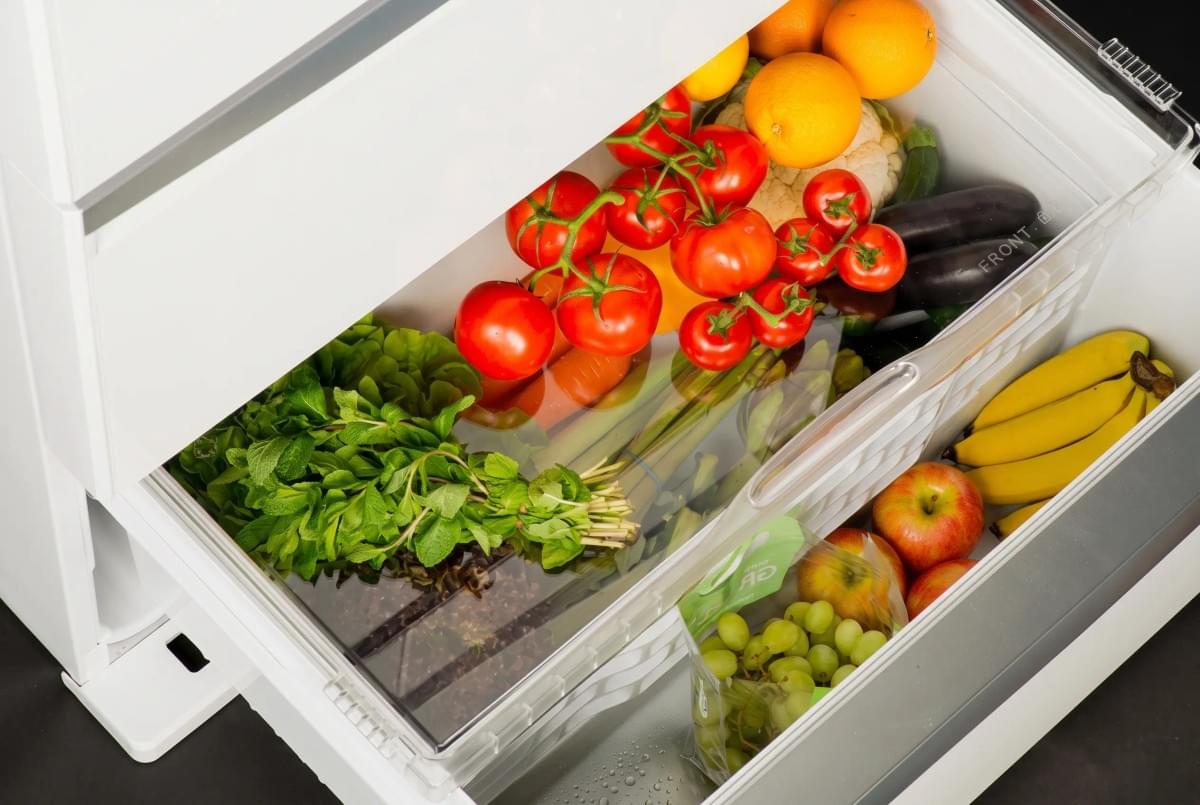 Top-down view into an open refrigerator crisp drawer filled with fresh tomatoes, citrus, bananas, and greens. Top-down view into an open refrigerator crisp drawer filled with fresh tomatoes, citrus, bananas, and greens.