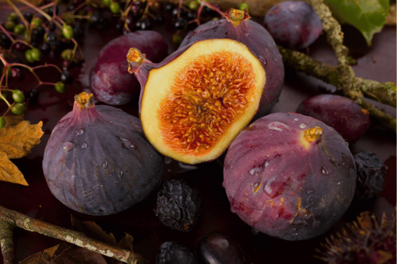 A moody, rustic arrangement of fresh figs, including a sliced half revealing golden-pink flesh, paired with dark autumn berries, dried fruits, and fallen leaves. A moody, rustic arrangement of fresh figs, including a sliced half revealing golden-pink flesh, paired with dark autumn berries, dried fruits, and fallen leaves.
