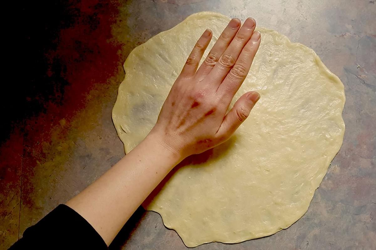 A hand presses down on a rolled-out pizza dough on a dark countertop. The dough is unevenly round, and the hand appears relaxed and focused. A hand presses down on a rolled-out pizza dough on a dark countertop. The dough is unevenly round, and the hand appears relaxed and focused.