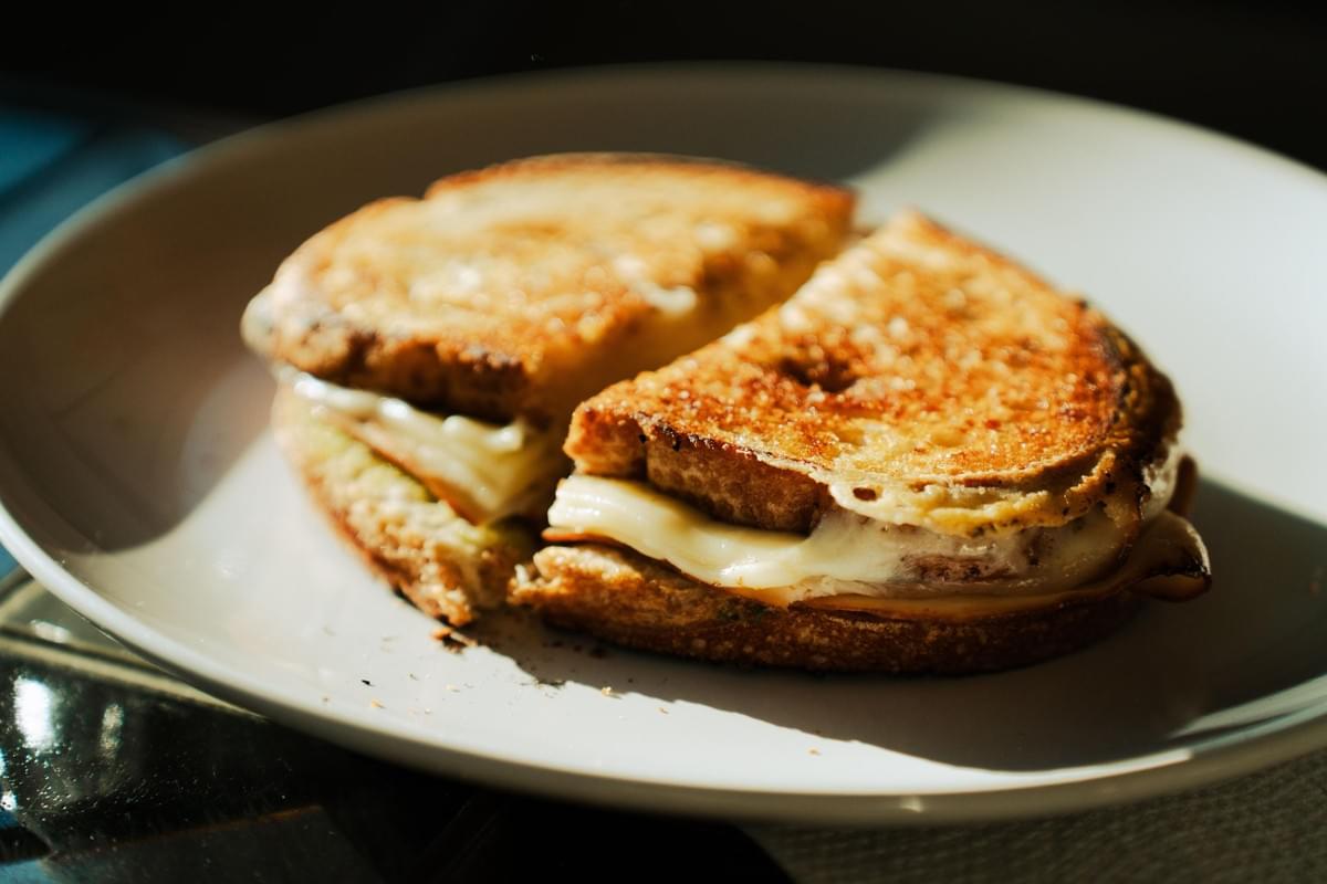 The image shows a golden-brown, toasted sandwich cut in half on a white plate. Melted white cheese and thin slices of meat ooze out from between the crispy, textured crusts under warm lighting. The image shows a golden-brown, toasted sandwich cut in half on a white plate. Melted white cheese and thin slices of meat ooze out from between the crispy, textured crusts under warm lighting.