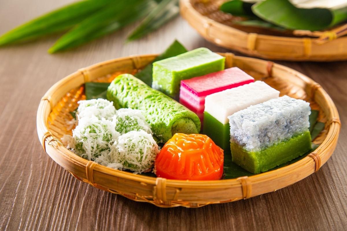A woven bamboo platter displays an assortment of colorful Peranakan or Malay Nyonya kuih, including green klepon coated in shredded coconut, layered pink and white rice cakes, and a bright orange Ang Ku Kueh. The traditional bite-sized desserts are arranged on a banana leaf liner against a warm wooden background. A woven bamboo platter displays an assortment of colorful Peranakan or Malay Nyonya kuih, including green klepon coated in shredded coconut, layered pink and white rice cakes, and a bright orange Ang Ku Kueh. The traditional bite-sized desserts are arranged on a banana leaf liner against a warm wooden background.
