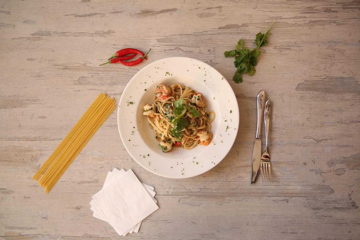 A top-down view shows a white bowl of cooked shrimp pasta garnished with fresh cilantro on a rustic wooden table. The plate is neatly surrounded by raw spaghetti, red chili peppers, fresh herbs, folded white napkins, and silver cutlery. A top-down view shows a white bowl of cooked shrimp pasta garnished with fresh cilantro on a rustic wooden table. The plate is neatly surrounded by raw spaghetti, red chili peppers, fresh herbs, folded white napkins, and silver cutlery.