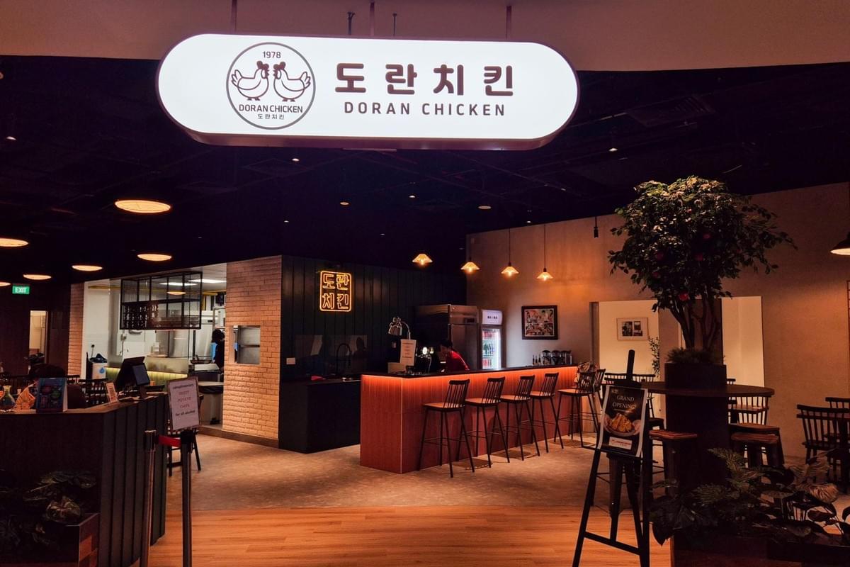 A glowing overhead sign reads "Doran Chicken" with Korean text and a two-chicken logo above a modern restaurant entrance. Inside, warm lighting illuminates a service counter with bar stools, an open kitchen area, and indoor plants. A glowing overhead sign reads "Doran Chicken" with Korean text and a two-chicken logo above a modern restaurant entrance. Inside, warm lighting illuminates a service counter with bar stools, an open kitchen area, and indoor plants.