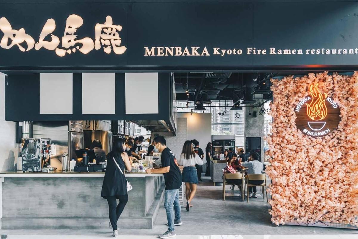 The storefront of "MENBAKA Kyoto Fire Ramen" features a modern black facade with Japanese calligraphy and English signage. Customers stand at the service counter while dining tables and a pink floral wall with a lit flame logo are visible in the background. The storefront of "MENBAKA Kyoto Fire Ramen" features a modern black facade with Japanese calligraphy and English signage. Customers stand at the service counter while dining tables and a pink floral wall with a lit flame logo are visible in the background.