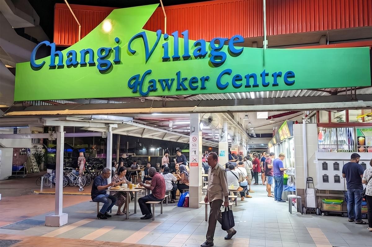 Changi Village Hawker Centre storefront with people dining and walking at night. Changi Village Hawker Centre storefront with people dining and walking at night.
