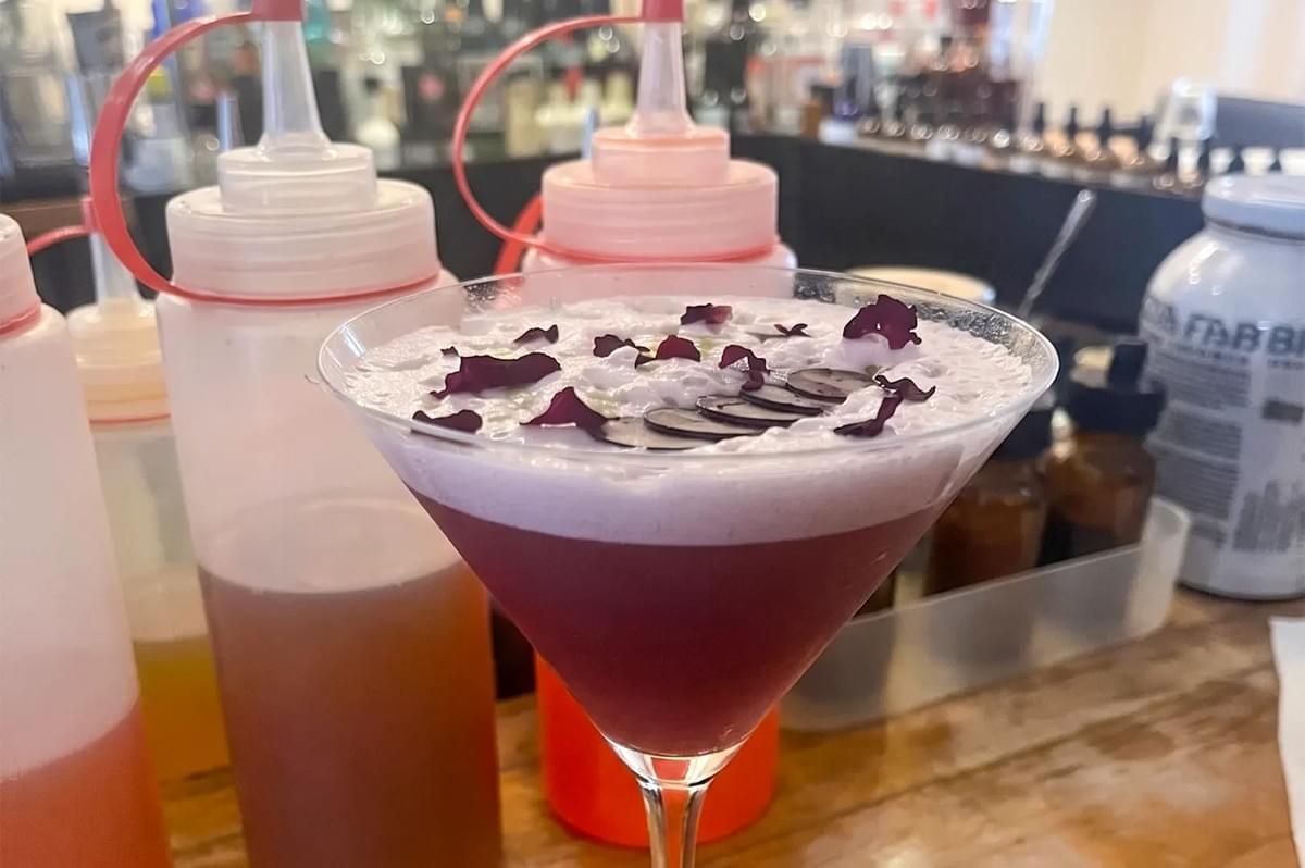A dark red cocktail in a martini glass topped with purple foam, sliced grapes, and dried flower petals. A dark red cocktail in a martini glass topped with purple foam, sliced grapes, and dried flower petals.