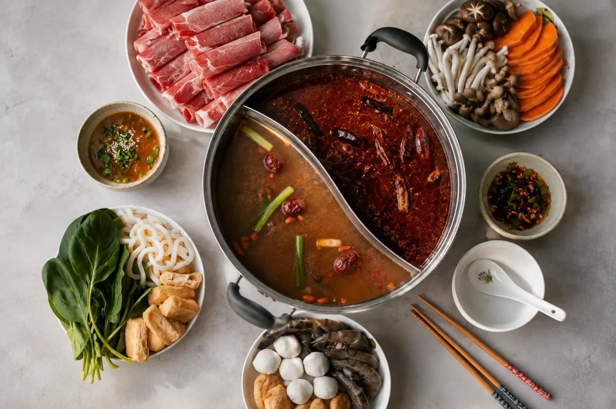 Overhead view of a split hot pot surrounded by raw sliced meat, vegetables, seafood, tofu, and dipping sauces. Overhead view of a split hot pot surrounded by raw sliced meat, vegetables, seafood, tofu, and dipping sauces.