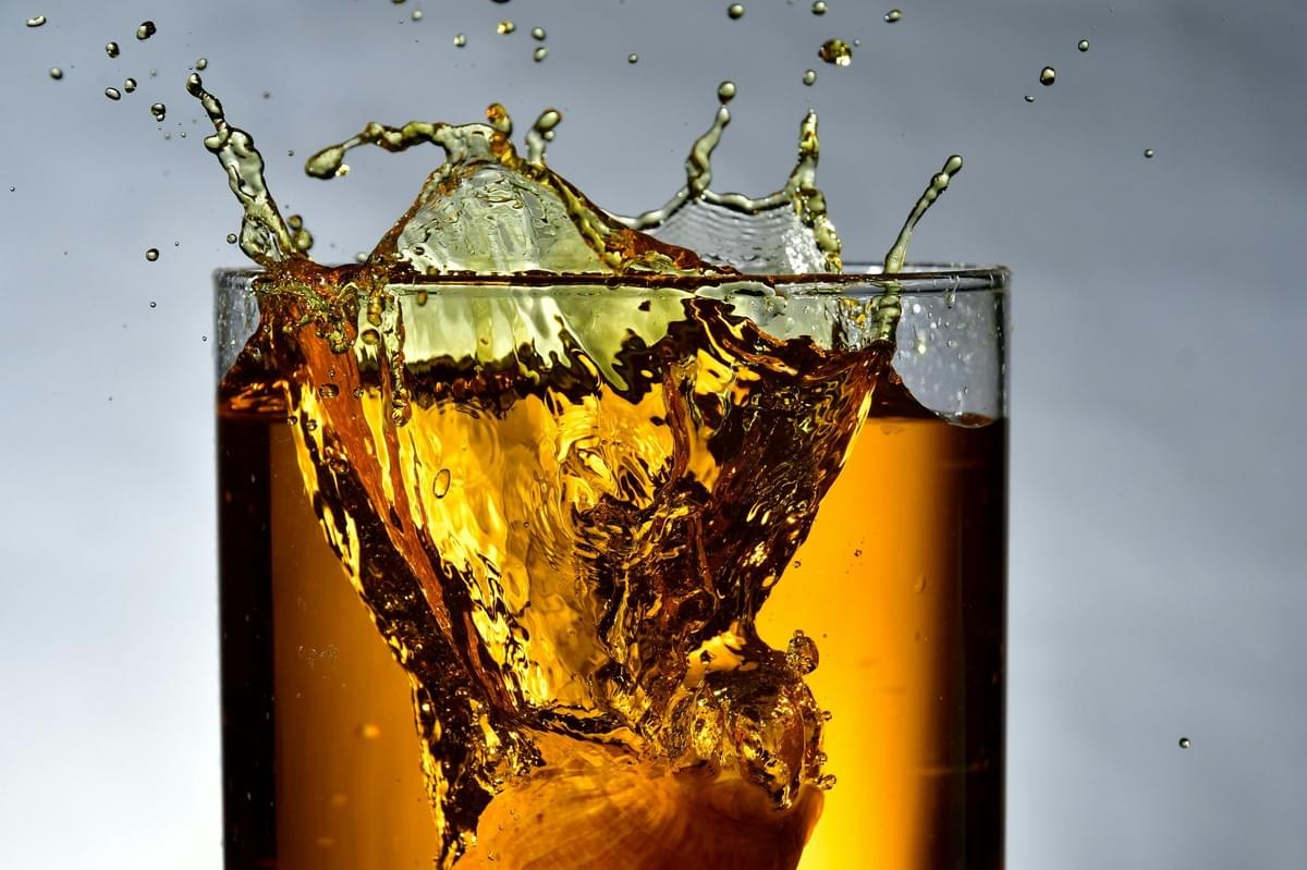 In the image, an object dropped into a clear glass creates a dramatic splash of amber-colored liquid. The liquid erupts over the rim in a crown-like wave, sending individual droplets flying against a neutral, grey background. In the image, an object dropped into a clear glass creates a dramatic splash of amber-colored liquid. The liquid erupts over the rim in a crown-like wave, sending individual droplets flying against a neutral, grey background.