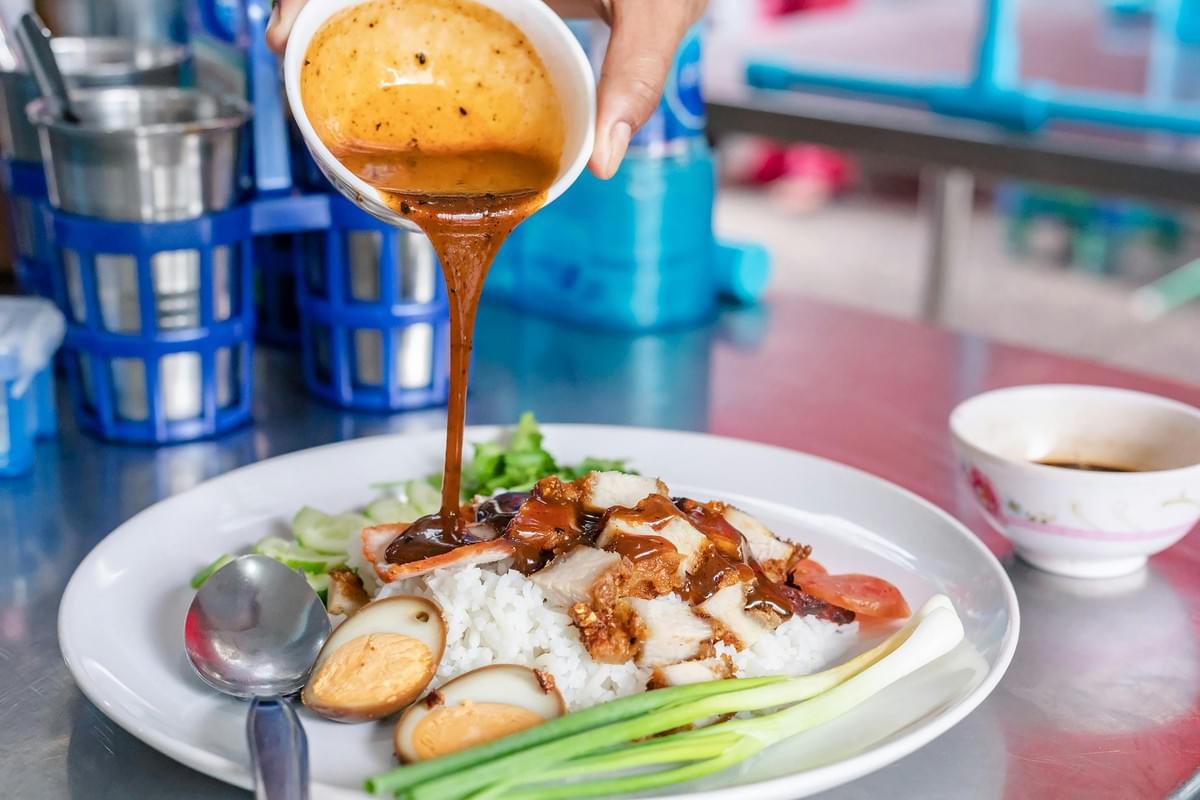 A person pours savory sauce from a small bowl over a plate of white rice topped with crispy pork belly, barbecue pork, and hard-boiled egg halves. The dish is served on a metal tabletop alongside fresh green onions, cucumbers, and traditional condiments. A person pours savory sauce from a small bowl over a plate of white rice topped with crispy pork belly, barbecue pork, and hard-boiled egg halves. The dish is served on a metal tabletop alongside fresh green onions, cucumbers, and traditional condiments.