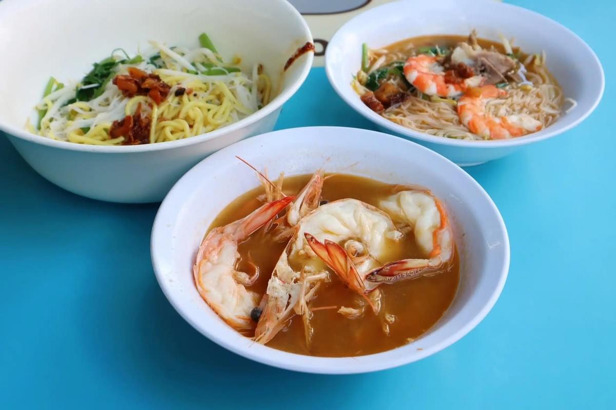 Three white bowls containing prawn noodles and soup are arranged on a blue tabletop. The foreground bowl features prominent large prawns in a rich broth, alongside bowls with noodles, greens, and additional toppings. Three white bowls containing prawn noodles and soup are arranged on a blue tabletop. The foreground bowl features prominent large prawns in a rich broth, alongside bowls with noodles, greens, and additional toppings.