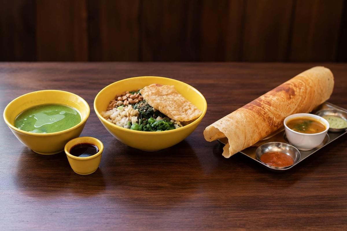 Wooden table featuring a yellow bowl of Thunder Tea Rice with greens and peanuts, a side of green herbal soup, and a crispy rolled dosa with chutneys Wooden table featuring a yellow bowl of Thunder Tea Rice with greens and peanuts, a side of green herbal soup, and a crispy rolled dosa with chutneys