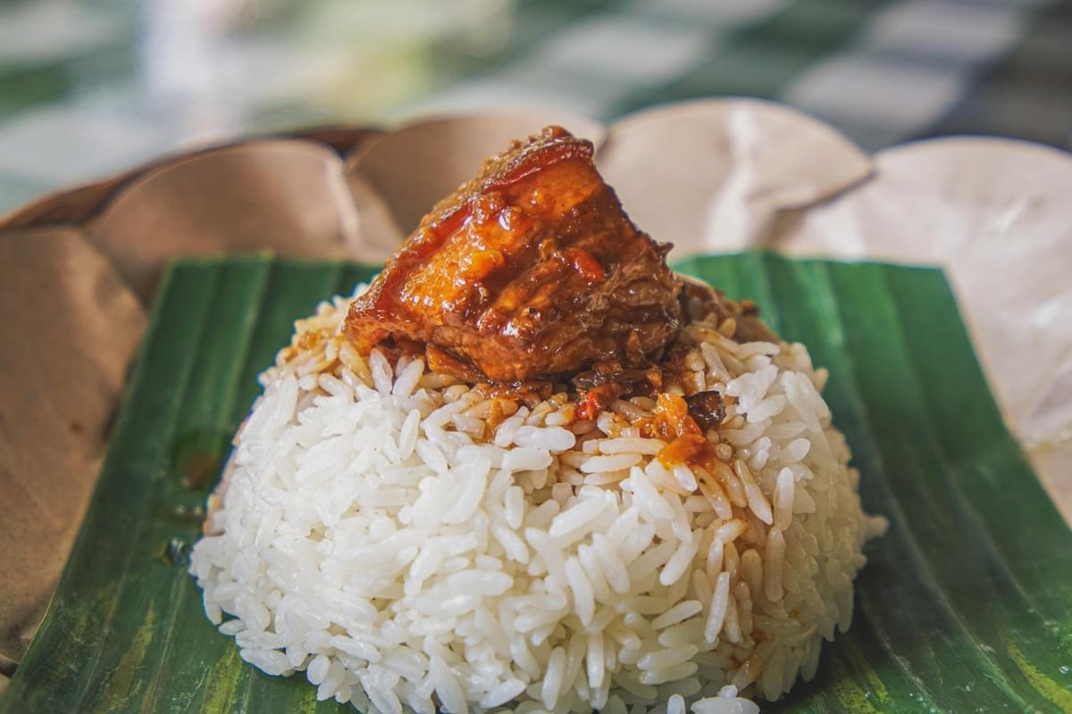 A mound of white rice sits on a green banana leaf, topped with a thick, savory piece of braised pork belly coated in a rich glaze. The dish is presented on a traditional woven plate with a blurred background. A mound of white rice sits on a green banana leaf, topped with a thick, savory piece of braised pork belly coated in a rich glaze. The dish is presented on a traditional woven plate with a blurred background.
