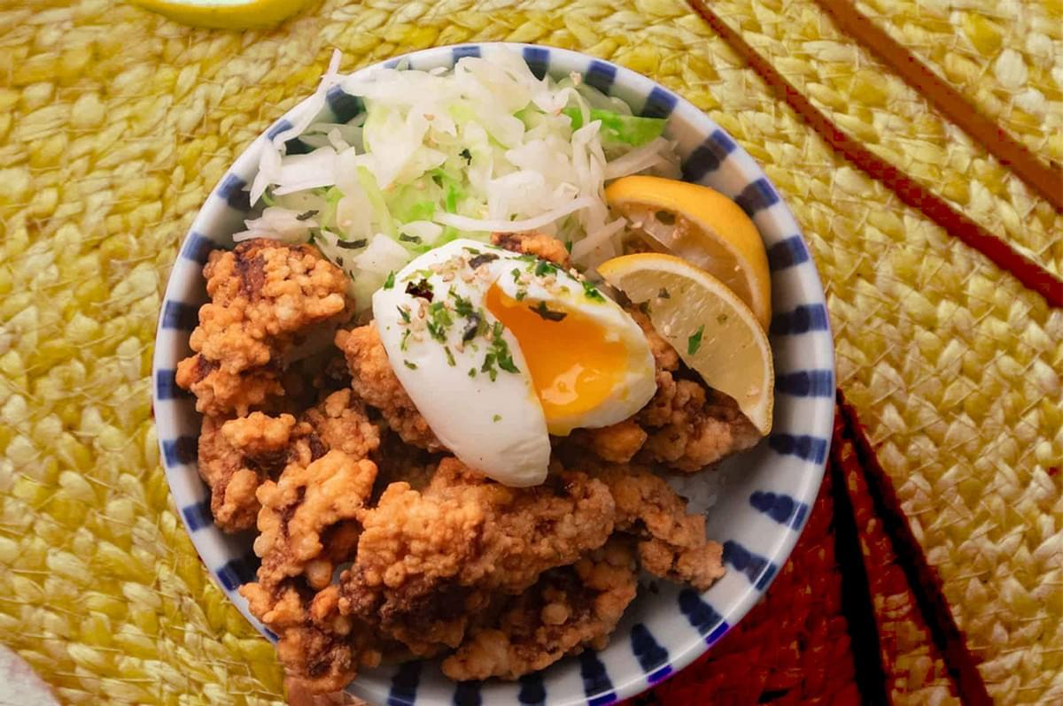 A bowl of crispy fried chicken topped with a soft-boiled egg, garnished with herbs, accompanied by shredded cabbage and lemon slices, on a woven mat. A bowl of crispy fried chicken topped with a soft-boiled egg, garnished with herbs, accompanied by shredded cabbage and lemon slices, on a woven mat.