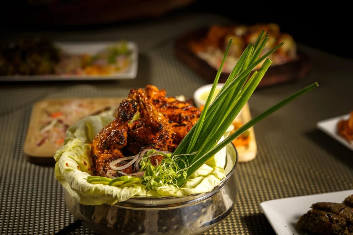 A metal bowl lined with cabbage leaves holds grilled, spiced chicken garnished with sliced onions and tall spring onion stalks. The dish is presented on a textured tablecloth with other blurred plates arranged in the background. A metal bowl lined with cabbage leaves holds grilled, spiced chicken garnished with sliced onions and tall spring onion stalks. The dish is presented on a textured tablecloth with other blurred plates arranged in the background.