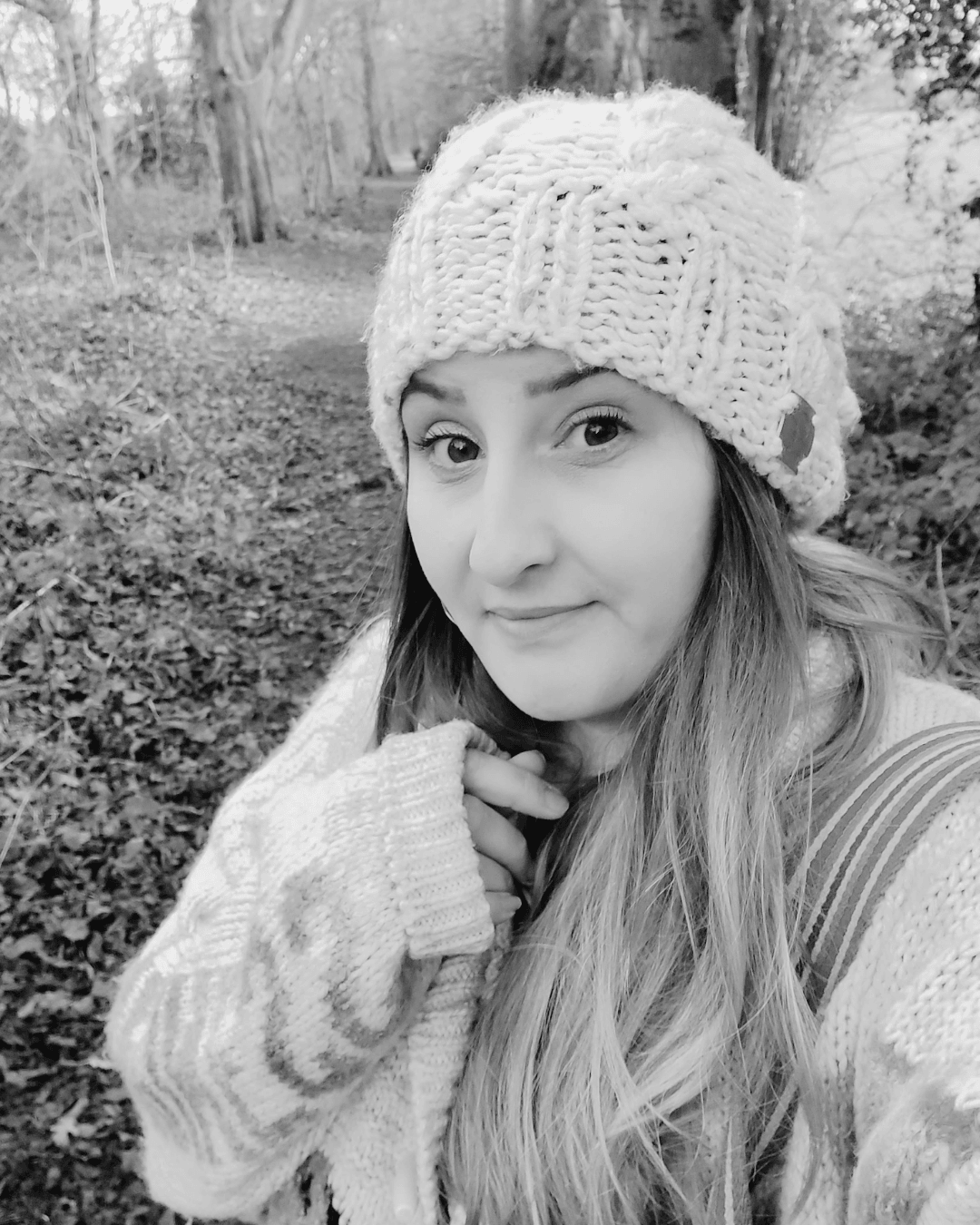 A black and white photograph of Charlie Edwards, a white woman in her thirties. Charlie stands in the woodland, with a leaf-covered path and trees behind her. She wears a light coloured jumper, pulled down over one hand, and a white woollen hat. Her brown hair falls around her shoulders. A black and white photograph of Charlie Edwards, a white woman in her thirties. Charlie stands in the woodland, with a leaf-covered path and trees behind her. She wears a light coloured jumper, pulled down over one hand, and a white woollen hat. Her brown hair falls around her shoulders.
