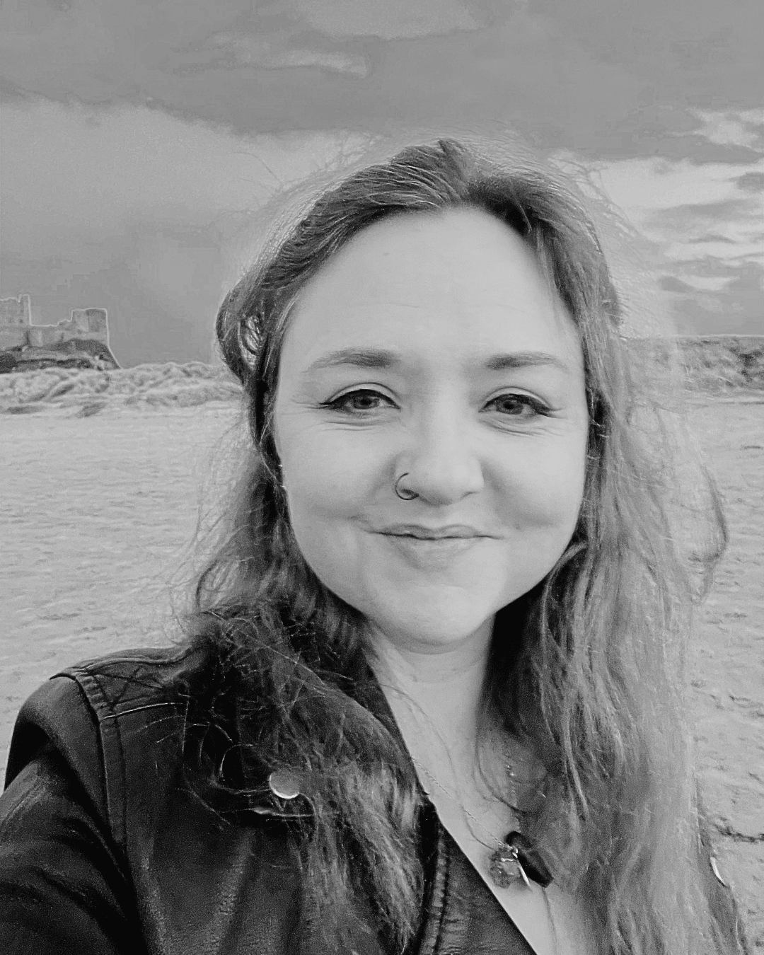 A black and white photograph of Ceryn Rowntree, a white woman in her 40s. Ceryn stands on a beach with the sky and a castle (Bamburgh in Northumberland) behind her. She wears a black jacket and has long, wavy dark hair over her shoulders. A black and white photograph of Ceryn Rowntree, a white woman in her 40s. Ceryn stands on a beach with the sky and a castle (Bamburgh in Northumberland) behind her. She wears a black jacket and has long, wavy dark hair over her shoulders.