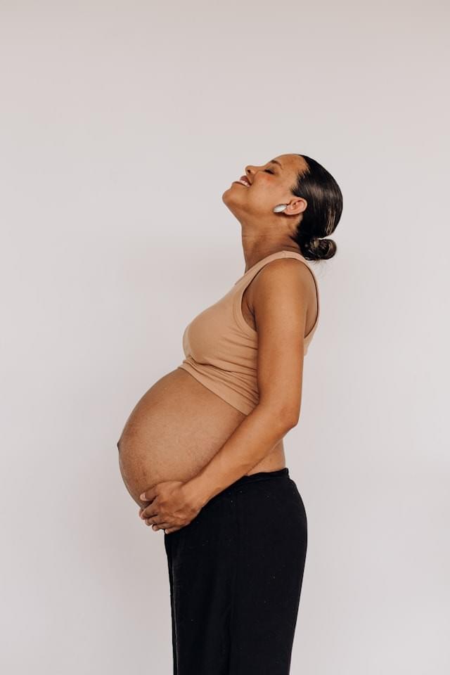 A third-trimester woman of colour holds her bump with both hands, smiling and looking upwards. A third-trimester woman of colour holds her bump with both hands, smiling and looking upwards.