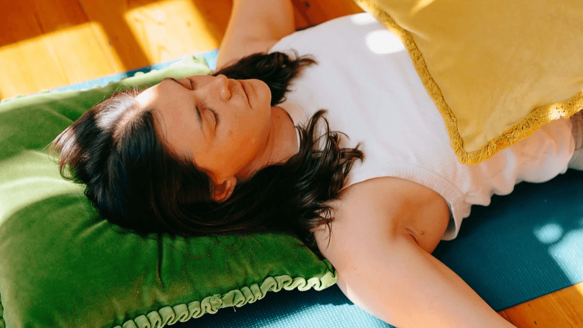 a woman relaxes on a yoga mat, her head on a green velvet cushion. Eyes closed, arms outstretched on the ground as sunlight pours onto the floor around her. a woman relaxes on a yoga mat, her head on a green velvet cushion. Eyes closed, arms outstretched on the ground as sunlight pours onto the floor around her.