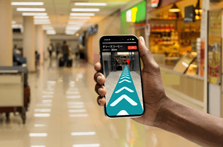 商業施設の通路で、手に持ったスマートフォン画面にARの進行方向ガイドが表示され、目的地までのルートを示している様子。 商業施設の通路で、手に持ったスマートフォン画面にARの進行方向ガイドが表示され、目的地までのルートを示している様子。