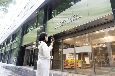 建物の入口前で白杖を持ち、スマートフォンを使って周囲を確認する視覚障害者の様子。 建物の入口前で白杖を持ち、スマートフォンを使って周囲を確認する視覚障害者の様子。