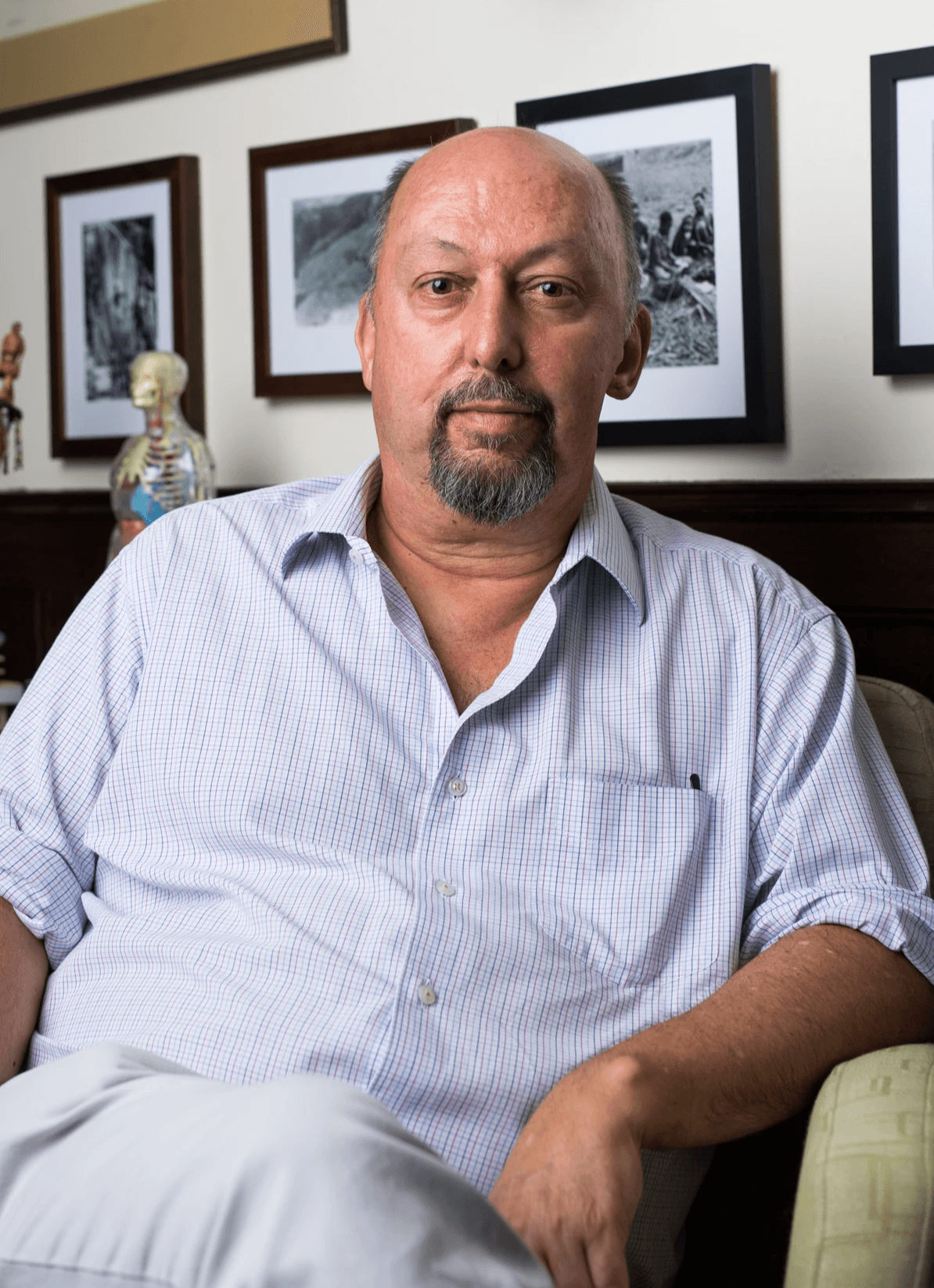 Portrait of a bearded man seated in front of black and white photographs, with a anatomical figure visible in the background Portrait of a bearded man seated in front of black and white photographs, with a anatomical figure visible in the background