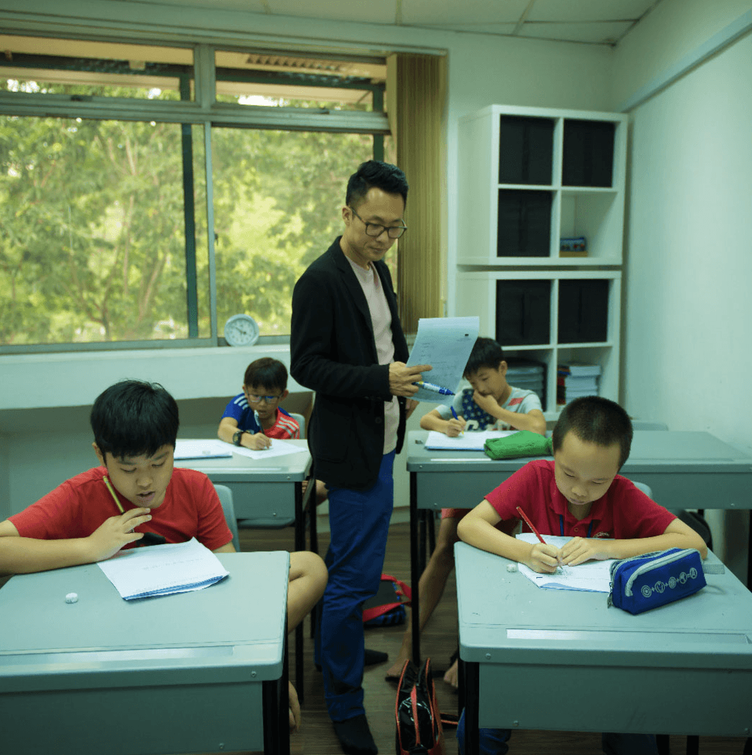 Teacher assisting four schoolchildren with written exercises in a bright classroom, with students seated at desks, worksheets and pencil cases on the tables, large windows overlooking greenery, and storage shelves in the background. Teacher assisting four schoolchildren with written exercises in a bright classroom, with students seated at desks, worksheets and pencil cases on the tables, large windows overlooking greenery, and storage shelves in the background.