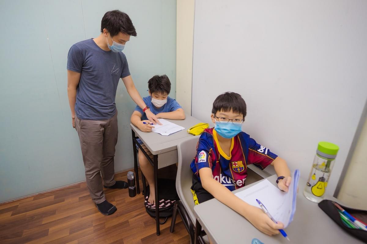 Teacher assisting two students wearing face masks while studying and writing at their desks in a classroom Teacher assisting two students wearing face masks while studying and writing at their desks in a classroom