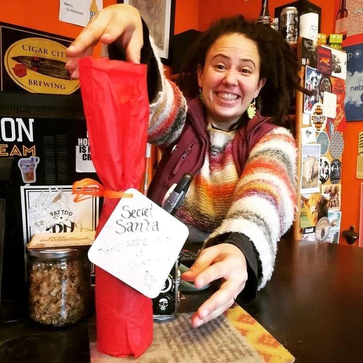 Jaclyn présentant une bouteille emballée sur le comptoir avec un petit mot accroché autour : Secret Santa. Remplacez-moi et emportez-moi ! Jaclyn présentant une bouteille emballée sur le comptoir avec un petit mot accroché autour : Secret Santa. Remplacez-moi et emportez-moi !