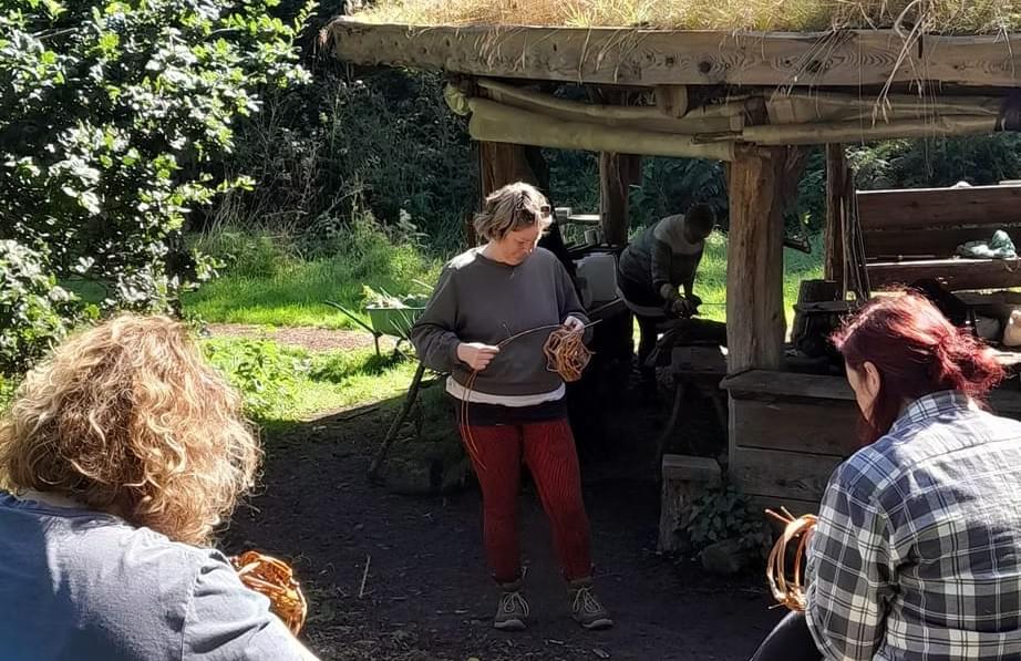 Three women are outside in a woodland setting, two have their backs turned away from the camera, one is facing forward but looking down towards a craft activity she is taking part in (similar to basket weaving). Three women are outside in a woodland setting, two have their backs turned away from the camera, one is facing forward but looking down towards a craft activity she is taking part in (similar to basket weaving).