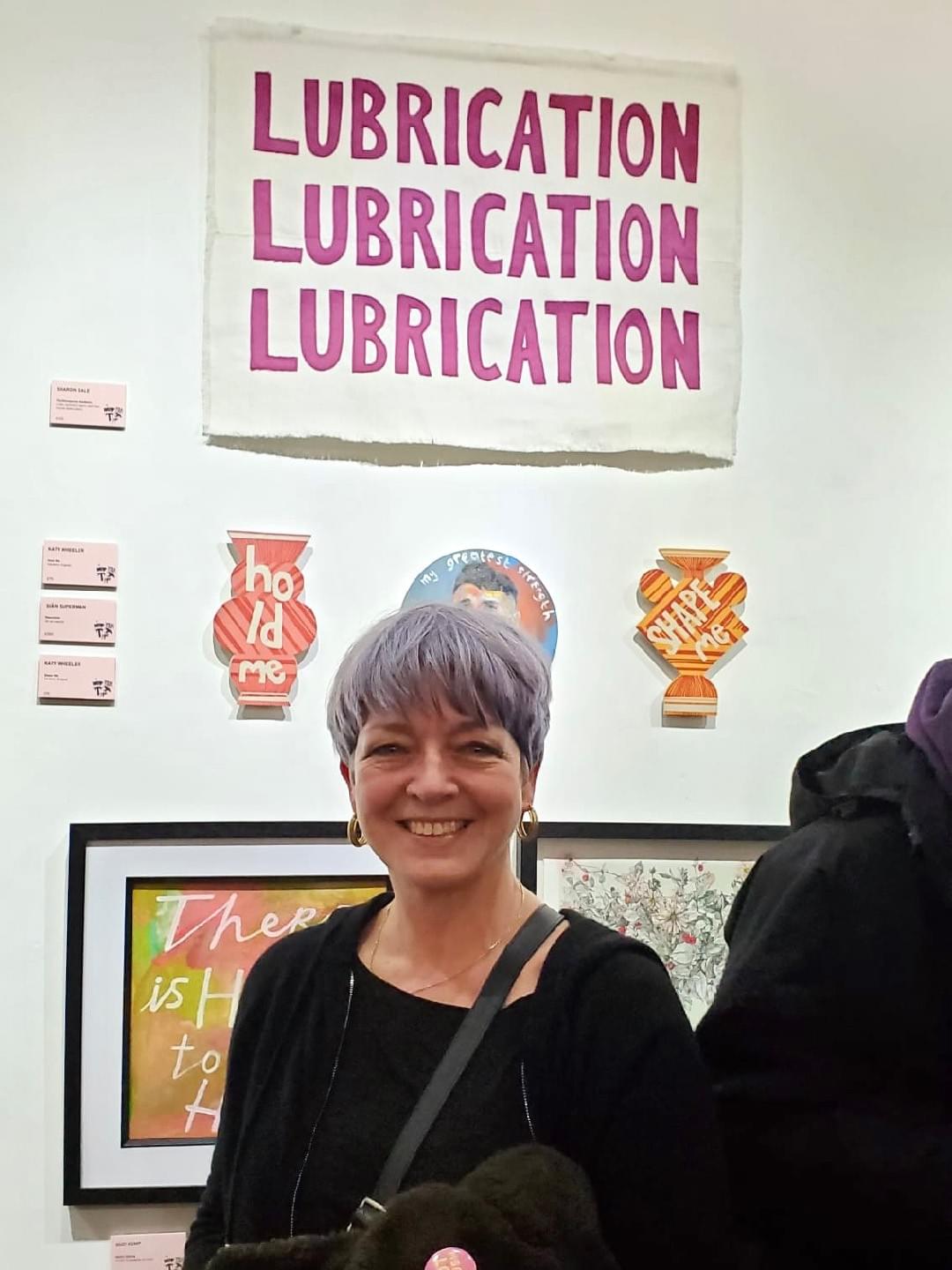 Head and shoulders shot of woman with short grey hair, wearing black. She is inside an art gallery with her back to a white wall with artworks hung behind her. Above her head is a textile artwork, pink fabric lettering is stitched onto cream linen. It says Lubrication, Lubrication, Lubrication. Head and shoulders shot of woman with short grey hair, wearing black. She is inside an art gallery with her back to a white wall with artworks hung behind her. Above her head is a textile artwork, pink fabric lettering is stitched onto cream linen. It says Lubrication, Lubrication, Lubrication.