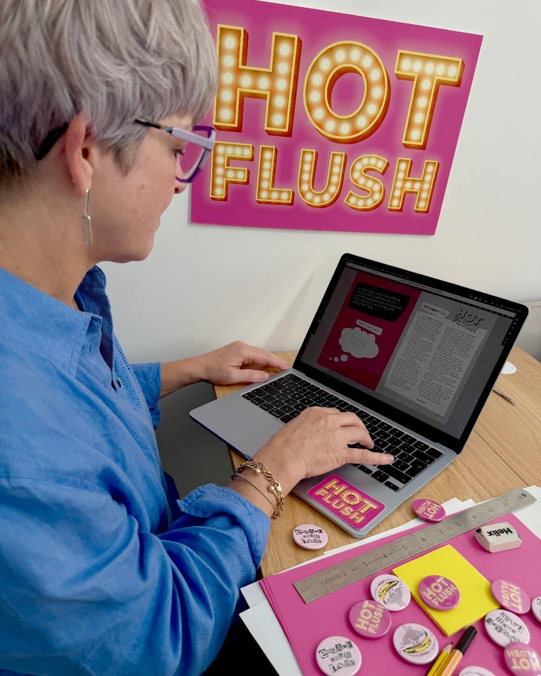 Woman, short grey hair, wearing glasses and a bright blue linen shirt. Sat at a desk with her laptop open. The zine artwork is visible on screen. In the background is a Hot Flush logo, pink background and yellow text. On the desk are papers in pink and white. Pen, pencil, rubber, ruler and lots of pin badges. Pin badges say Hot Flush; pink background and yellow text. Engage Your Rage; pale pink background, black and white ransom style text. A pop art style illustrated banana in yellow and black on a pale pink background. Woman, short grey hair, wearing glasses and a bright blue linen shirt. Sat at a desk with her laptop open. The zine artwork is visible on screen. In the background is a Hot Flush logo, pink background and yellow text. On the desk are papers in pink and white. Pen, pencil, rubber, ruler and lots of pin badges. Pin badges say Hot Flush; pink background and yellow text. Engage Your Rage; pale pink background, black and white ransom style text. A pop art style illustrated banana in yellow and black on a pale pink background.