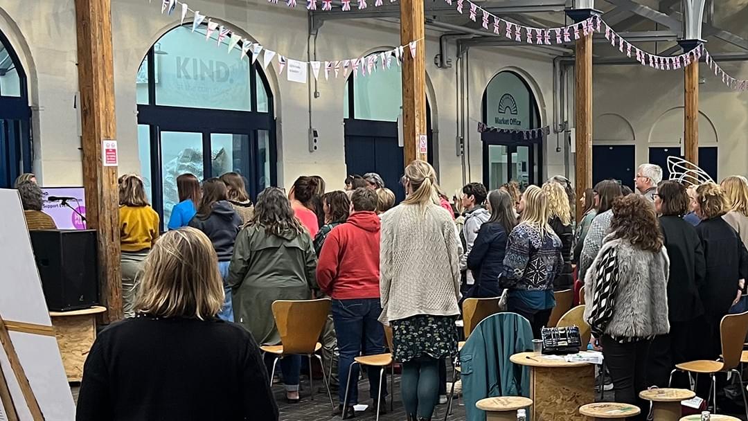 People, mainly women, are standing with their backs to the camera. They are listening to a woman speaking into a microphone People, mainly women, are standing with their backs to the camera. They are listening to a woman speaking into a microphone