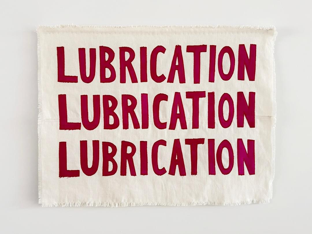 Hanging textle artwork on a white wall. Rectangular cream linen, frayed edges, with bright pink, sewn lettering which say Lubrication, Lubrication, Lubrication, stacked above one another. Hanging textle artwork on a white wall. Rectangular cream linen, frayed edges, with bright pink, sewn lettering which say Lubrication, Lubrication, Lubrication, stacked above one another.