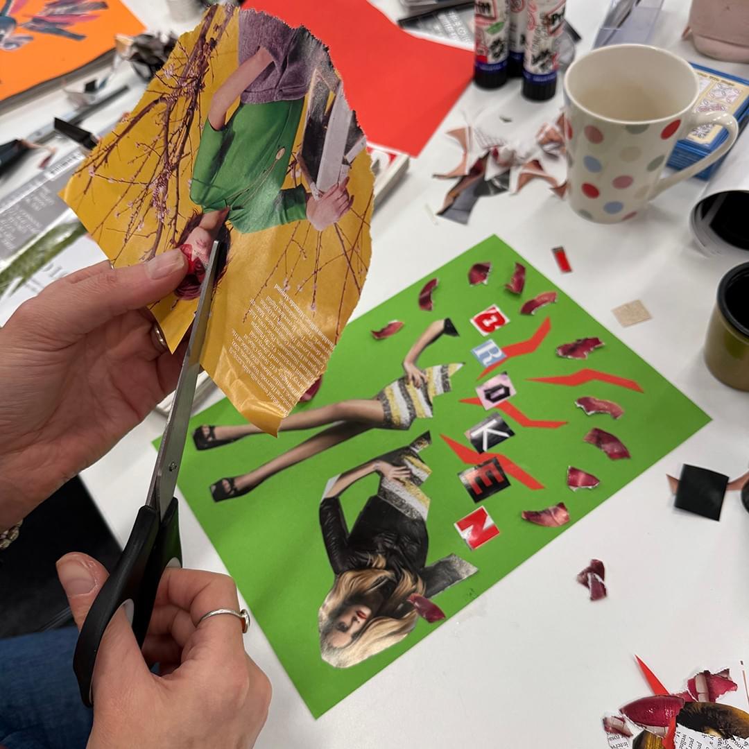A close-up shot of hands cutting an image from a magazine. In the background is the table with a collage artwork in progress, glue sticks, and a cup of tea. A close-up shot of hands cutting an image from a magazine. In the background is the table with a collage artwork in progress, glue sticks, and a cup of tea.