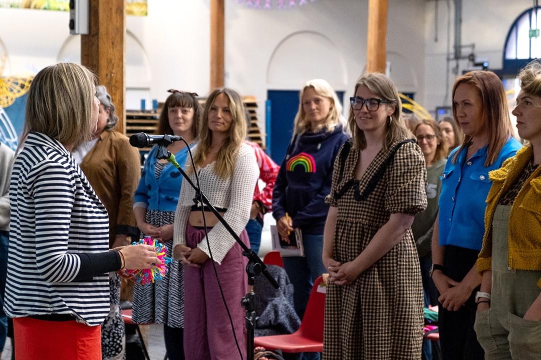 A group of women standing up, and facing towards a woman who is speaking into a microphone A group of women standing up, and facing towards a woman who is speaking into a microphone