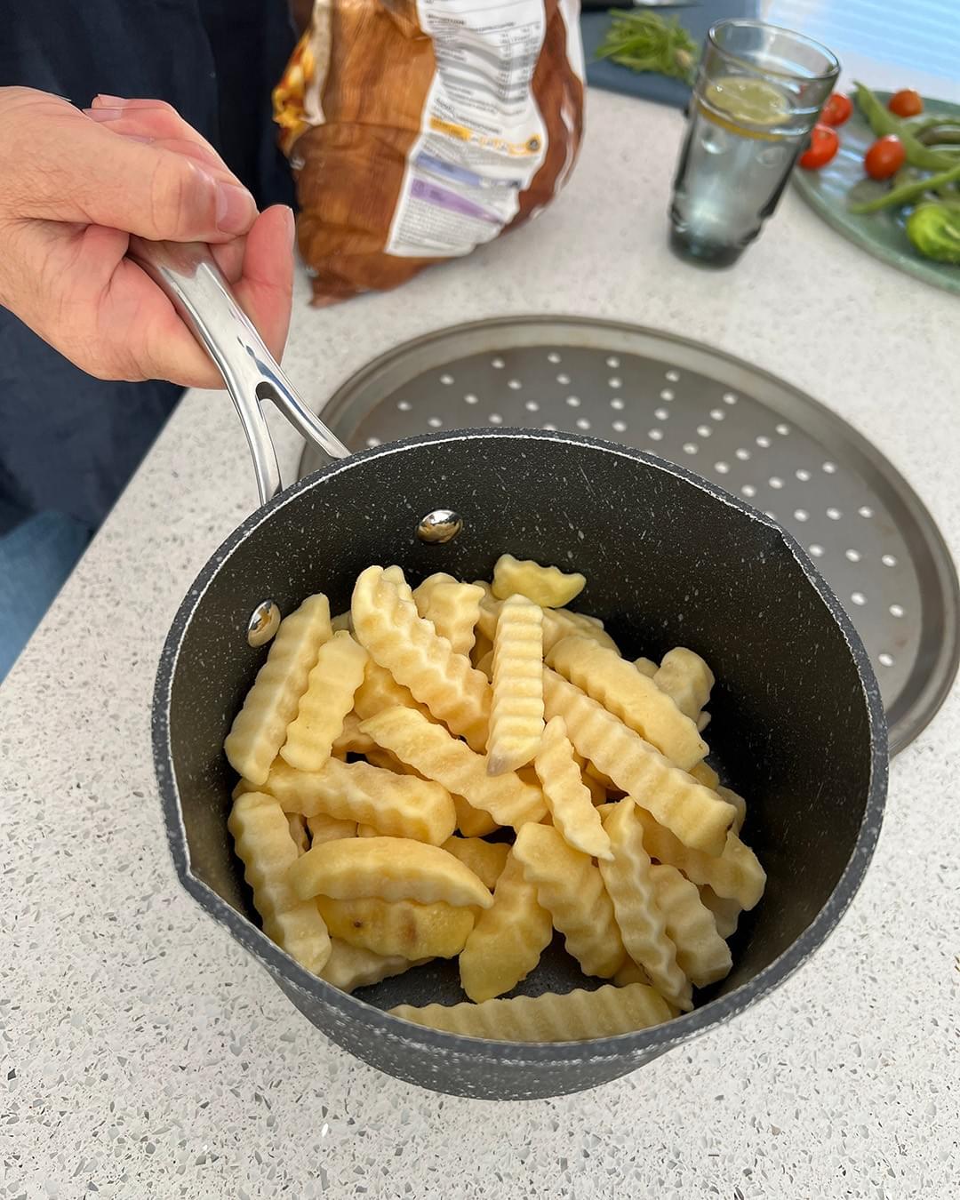 A hand is holding a saucepan which is full with oven chips. Just behind the saucepan is an oven baking tray. In the background is a bag of frozen oven chips, a glass of water with a slice of lemon, and a flat dish of tomatoes and runners beans. A hand is holding a saucepan which is full with oven chips. Just behind the saucepan is an oven baking tray. In the background is a bag of frozen oven chips, a glass of water with a slice of lemon, and a flat dish of tomatoes and runners beans.