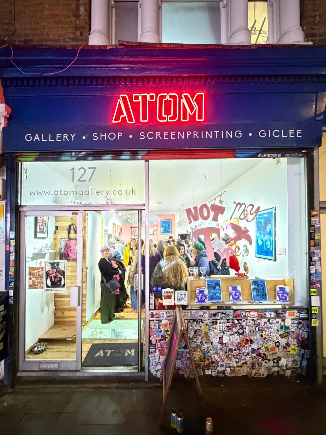 Outside Atom Gallery looking in. Brighly lit, you can view inside the windows. It is busy with people chatting and mingling. Atom’s sign is an electric light in bright red. Not My Type is painted in pink in the window. The gallery’s exterior wall is plastered with stickers. Through the windows, artwork can be seen hanging on the white and pink walls. Outside Atom Gallery looking in. Brighly lit, you can view inside the windows. It is busy with people chatting and mingling. Atom’s sign is an electric light in bright red. Not My Type is painted in pink in the window. The gallery’s exterior wall is plastered with stickers. Through the windows, artwork can be seen hanging on the white and pink walls.