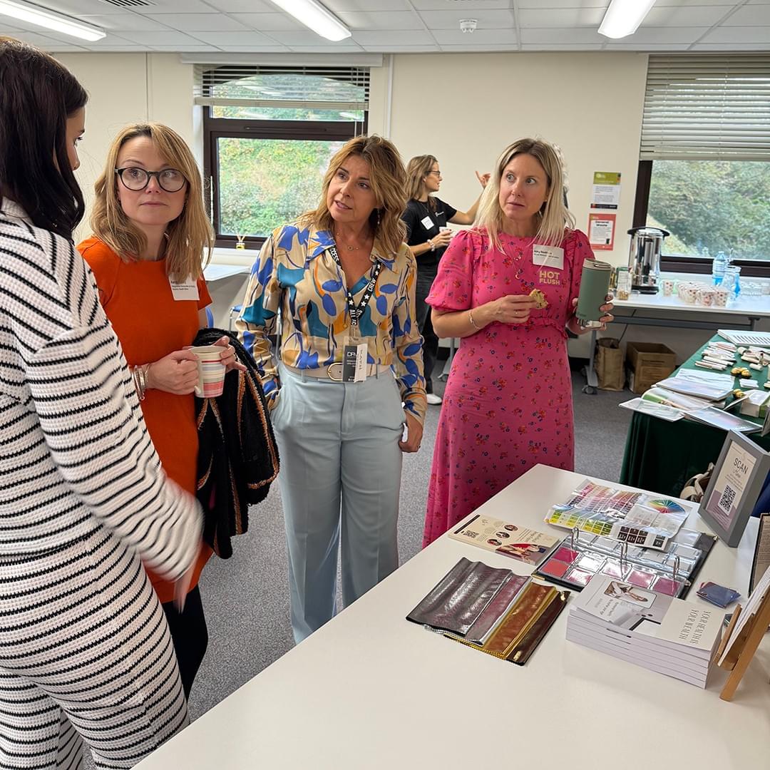 Four women are standing by a table and are chatting together. On the table is a display of books, fabric samples, and colour charts. Four women are standing by a table and are chatting together. On the table is a display of books, fabric samples, and colour charts.