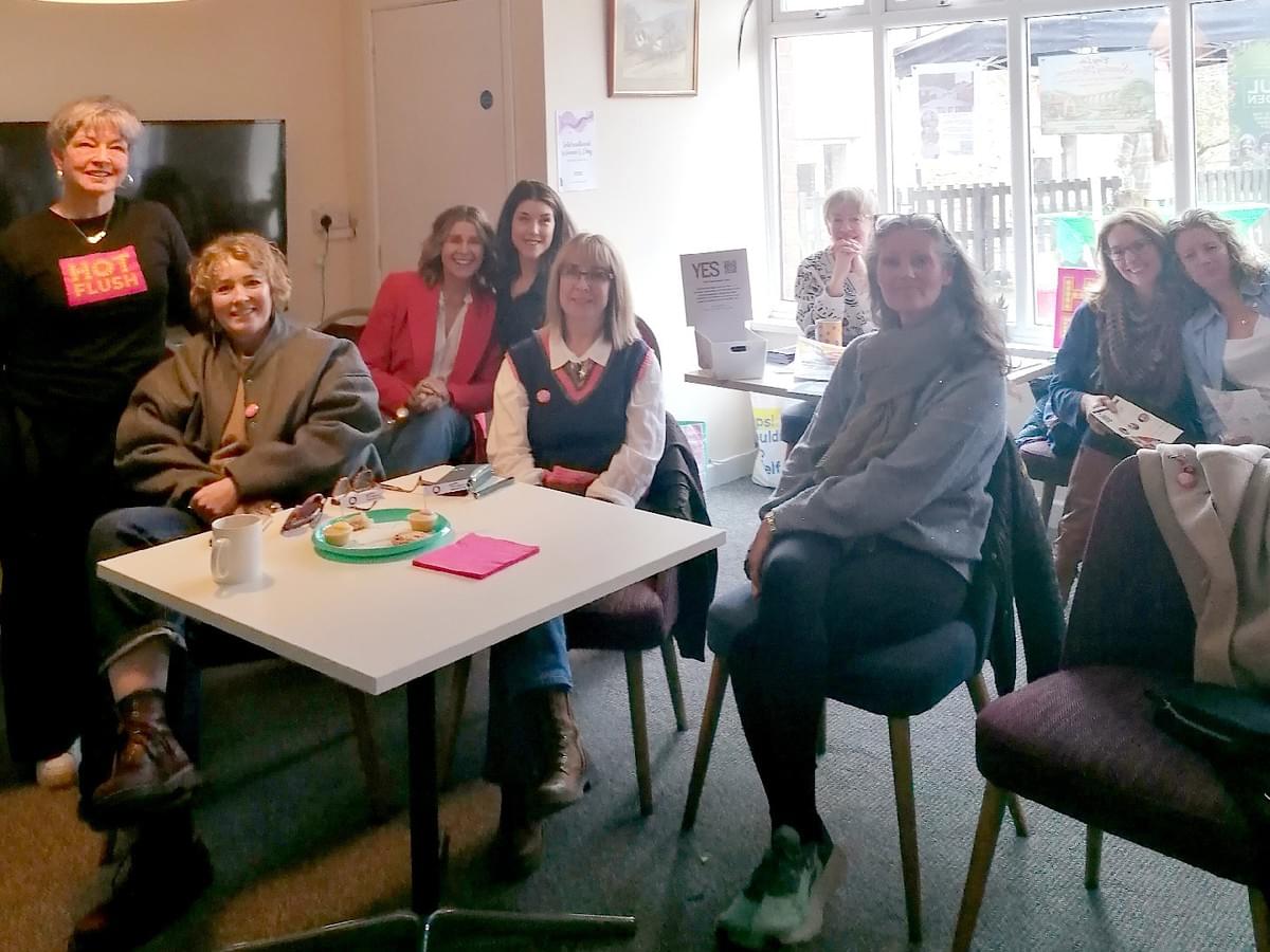 A group of women standing up, and facing towards a woman who is speaking into a microphone A group of women standing up, and facing towards a woman who is speaking into a microphone