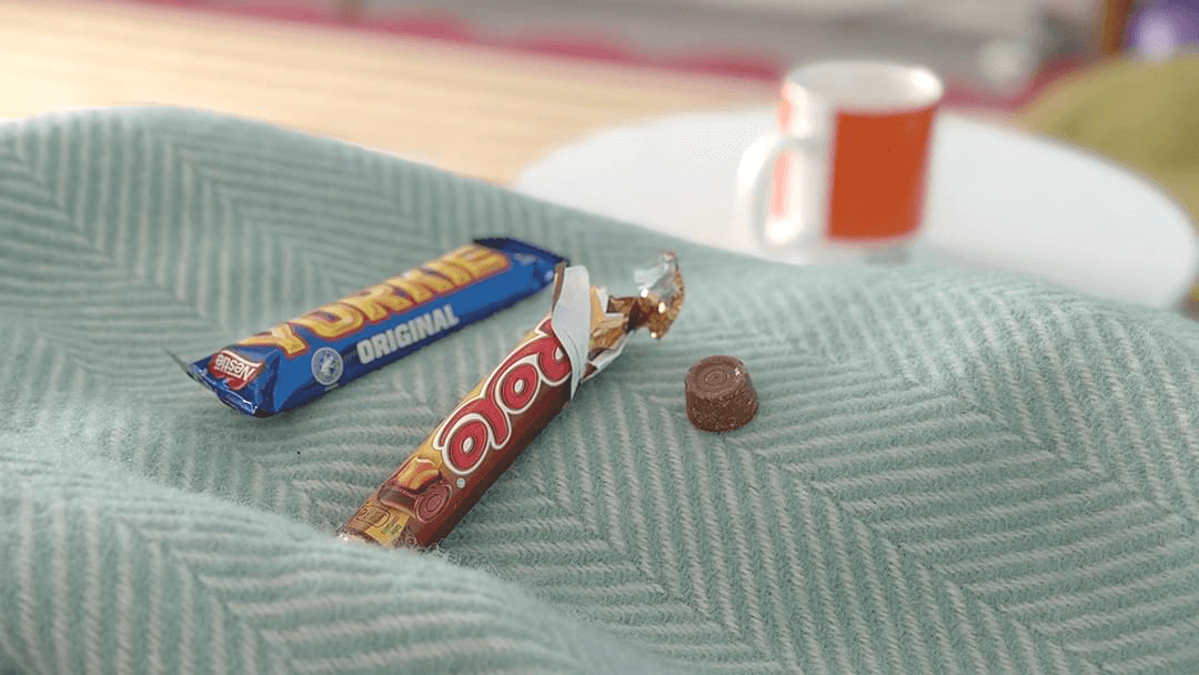 The setting is a lounge. Two chocolate bars, a Yorkie and packet of Rolos are placed on a blue and white herringbone patterned blanket. One Rolo has been taken out of its packet and placed slightly to the right. The background is blurred, but there is a small white table and on it is an orange and white mug. The setting is a lounge. Two chocolate bars, a Yorkie and packet of Rolos are placed on a blue and white herringbone patterned blanket. One Rolo has been taken out of its packet and placed slightly to the right. The background is blurred, but there is a small white table and on it is an orange and white mug.